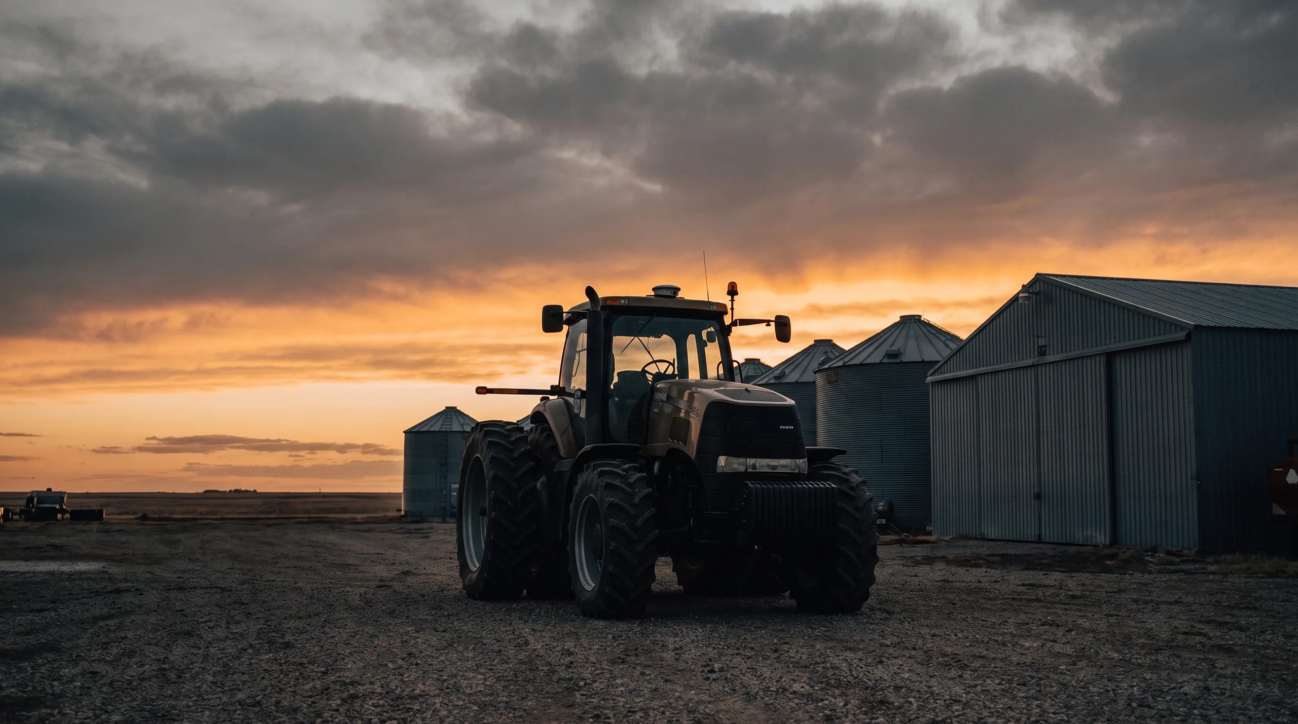 A row-crop tractor on dual wheels parked in a gravel farmyard under a prairie sunset, a Northing receiver mounted small on its cab roof, grain bins and a steel shed behind it
